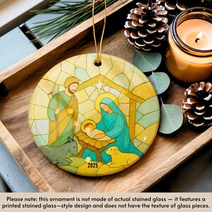 May include: A round Christmas ornament with a stained-glass style design depicting the Nativity scene. The ornament features Joseph, Mary, and baby Jesus, with a donkey and a lamb. The year "2025" is printed on the ornament. The ornament is displayed on a wooden tray with pine cones and a lit candle.