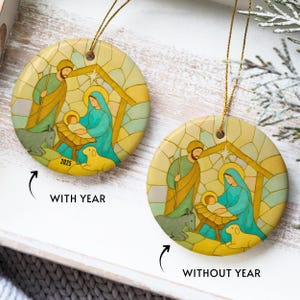 May include: Round Christmas ornament with a stained-glass style Nativity scene. The design features Mary, Joseph, and baby Jesus in a manger, with a donkey and sheep. The ornament has a gold string for hanging and the year 2025 is printed on one of the ornaments.