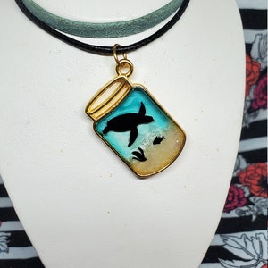 Puede incluir: Un collar con colgante dorado con una silueta de tortuga dentro de un tarro con un fondo azul y arena. El collar tiene un cordón negro y turquesa.