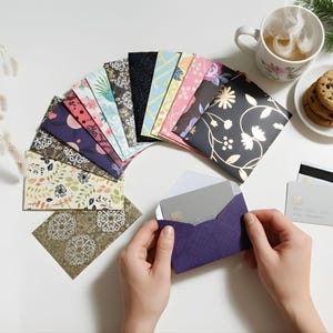 May include: A collection of patterned envelopes in various colors and designs, arranged in a fan shape. A person is holding a purple envelope with a card inside. A cup of tea, cookies, and credit cards are also present.