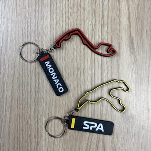 May include: Two keyrings with race track outlines. One has a red outline and a black tag with "MONACO" in white. The other has a yellow outline and a black tag with "SPA" in white.