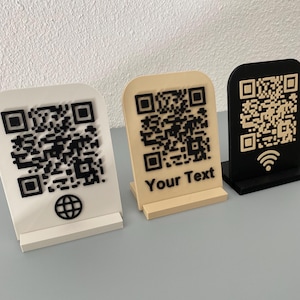 May include: Three QR code stands in white, beige, and black. Each stand has a unique QR code design. The beige stand has the text "Your Text" at the bottom. The white stand has a globe icon, and the black stand has a wifi icon.