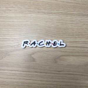 Könnte beinhalten: Ein weißer Aufkleber mit dem Namen "RACHEL" in Schwarz, im Stil der Fernsehserie "Friends". Die Buchstaben haben farbige Punkte darüber. Der Aufkleber liegt auf einer hellbraunen Holzoberfläche.