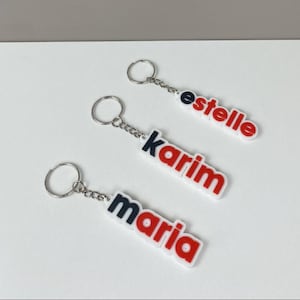 May include: Three personalized keychains with names: "estelle", "karim", and "maria". Each keychain features a white base with red and black lettering, attached to a silver keyring. The keychains are arranged on a white surface.