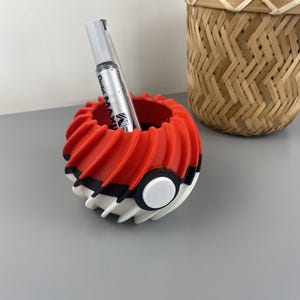 Peut inclure: Un organisateur de bureau sphérique présentant un motif en spirale avec des nervures rouges, blanches et noires. L'objet comporte un bouton circulaire blanc au centre et contient un marqueur argenté Pentel Maxiflo dans sa partie supérieure.
