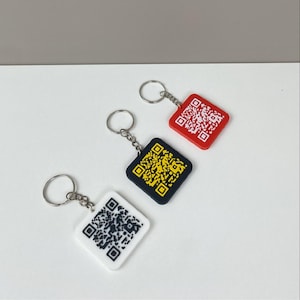 Op de afbeelding: Drie sleutelhangers met QR-codes. Eén sleutelhanger is rood met een witte QR-code, één is zwart met een gele QR-code en één is wit met een zwarte QR-code. Elke sleutelhanger heeft een zilveren sleutelring.