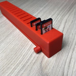 Peut inclure: Un support de cartouche de jeu rouge vif pour Nintendo Switch. Le support comporte des fentes pour plusieurs cartouches de jeu et un petit bouton. Trois cartouches de jeu sont insérées. Le support est en plastique.