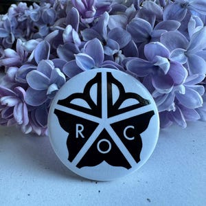May include: A white button with a black star-shaped design and the letters "R", "O", and "C". The button is set against a backdrop of purple flowers, creating a visually appealing contrast. The button's design is geometric and eye-catching.