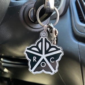 May include: A black and white keychain with a flower-shaped design and the letters "ROC" in the center. The keychain is attached to a silver key ring with keys.