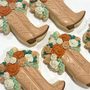 May include: Several cowboy boot-shaped cookies decorated with tan icing and floral arrangements. The boots have decorative swirls and are adorned with orange, white, and green frosting flowers. The cookies are arranged on a white surface.