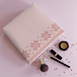 May include: A light pink cosmetic bag with a snowflake pattern, surrounded by makeup items like a brush, mascara, and eyeshadow. The bag is rectangular and has a knitted texture. Scattered confetti adds a touch of sparkle.