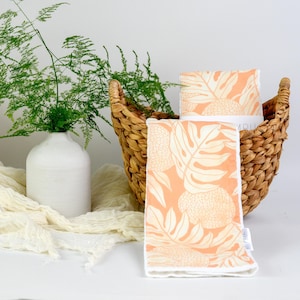 May include: A rectangular cloth featuring a peach-colored background with a white botanical leaf and fruit pattern. The cloth has a white border and is folded, with one piece placed inside a woven brown basket and another draped in front.