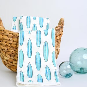 May include: White cloth napkins with a blue surfboard pattern are displayed in a woven basket. Two clear glass spheres are next to the basket. The napkins have a repeating pattern of blue surfboards.
