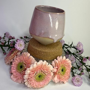 Könnte beinhalten: Eine helllilafarbene Keramikschale mit texturierter Glasur steht auf einem Korkuntersetzer, umgeben von rosa und violetten Blumen.