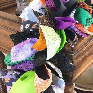 May include: A decorative garland constructed from numerous tied fabric strips in various textures and patterns. The colors include bright lime green, deep purple, orange, and black, featuring prints of pumpkins, swirls, and glittery accents.