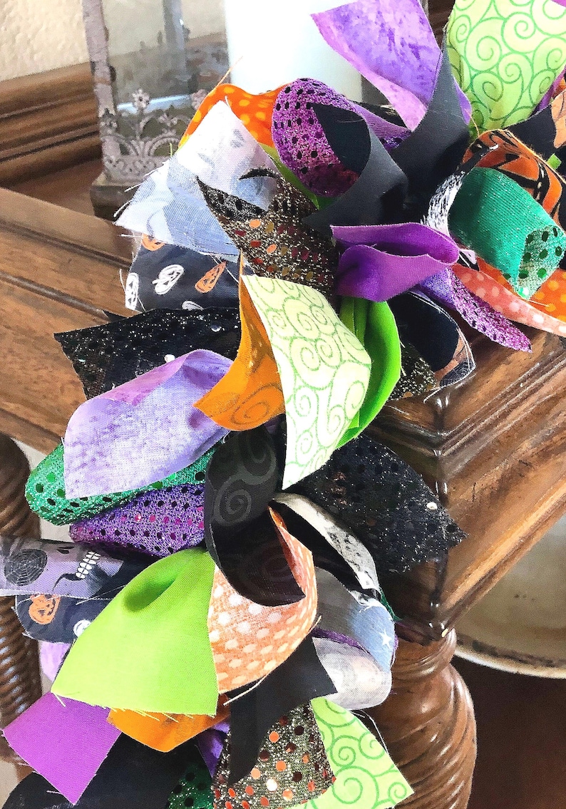 May include: A decorative garland constructed from numerous tied fabric strips in various textures and patterns. The colors include bright lime green, deep purple, orange, and black, featuring prints of pumpkins, swirls, and glittery accents.