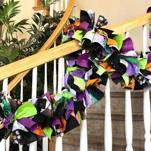 May include: A Halloween garland made of fabric scraps in black, green, orange, and purple. The garland is draped over a white banister and is decorated with various Halloween-themed patterns.