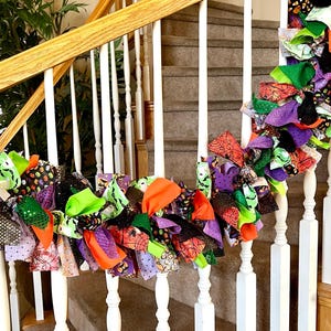 May include: A decorative fabric garland featuring a mix of knotted strips in orange, purple, green, and black. The fabric patterns include spider webs, pumpkins, and polka dots, draped along a white wooden staircase railing with vertical balusters.
