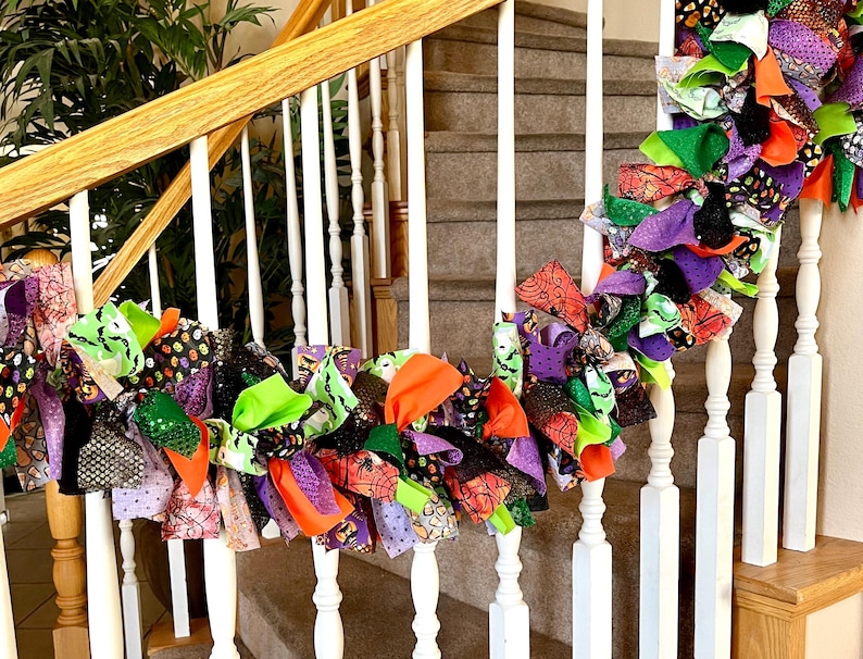 May include: A decorative fabric garland featuring a mix of knotted strips in orange, purple, green, and black. The fabric patterns include spider webs, pumpkins, and polka dots, draped along a white wooden staircase railing with vertical balusters.