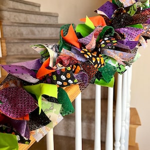 May include: A decorative Halloween garland made of tied fabric strips in various textures and patterns, including purple, orange, lime green, and black. The fabric features prints of pumpkins, candy corn, ghosts, and bats with glittery accents.