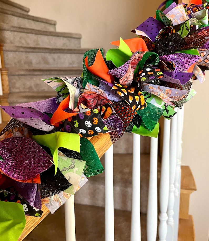 May include: A decorative Halloween garland made of tied fabric strips in various textures and patterns, including purple, orange, lime green, and black. The fabric features prints of pumpkins, candy corn, ghosts, and bats with glittery accents.