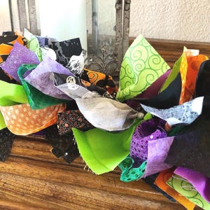 May include: A decorative fabric garland featuring a mix of knotted fabric strips in vibrant shades of orange, lime green, purple, and black. The fabric pieces include various patterns such as polka dots, swirls, and solid colors, creating a textured, layered appearance throughout the length of the garland.