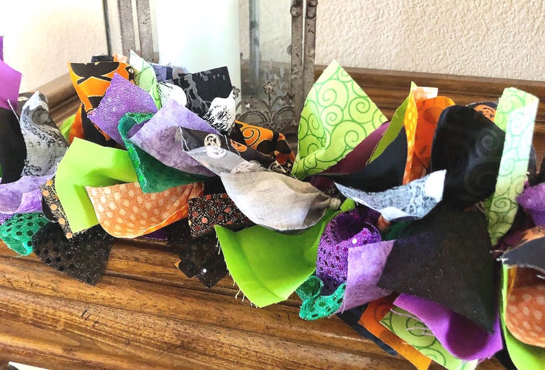 May include: A decorative fabric garland featuring a mix of knotted fabric strips in vibrant shades of orange, lime green, purple, and black. The fabric pieces include various patterns such as polka dots, swirls, and solid colors, creating a textured, layered appearance throughout the length of the garland.