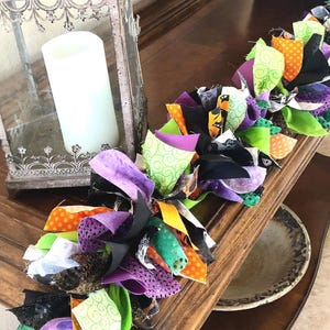 May include: A decorative fabric rag garland featuring a mix of black, purple, lime green, and orange fabric strips with various patterns including polka dots and swirls, arranged in a knotted design alongside a metal lantern with a white candle.
