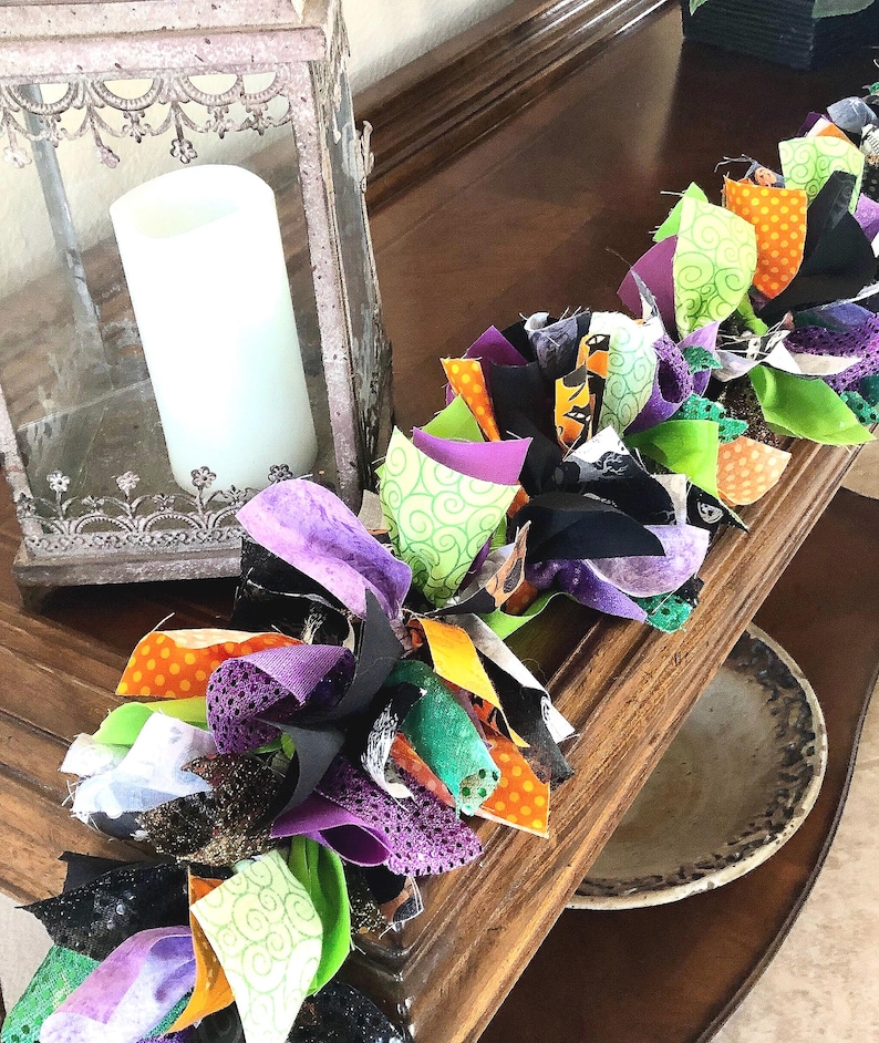 May include: A decorative fabric rag garland featuring a mix of black, purple, lime green, and orange fabric strips with various patterns including polka dots and swirls, arranged in a knotted design alongside a metal lantern with a white candle.