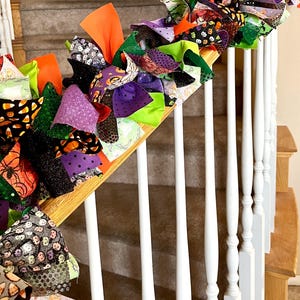 May include: A decorative fabric rag garland featuring a mix of orange, purple, green, and black fabric strips. The garland includes various patterns such as candy corn, pumpkins, spiders, and polka dots, tied together to create a textured display.