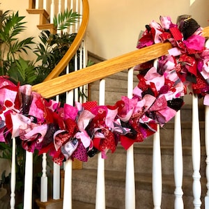 May include: A pink, red, and black fabric garland with hearts and other patterns hangs on a white railing. The garland is made of scraps of fabric and is perfect for Valentine's Day decor.