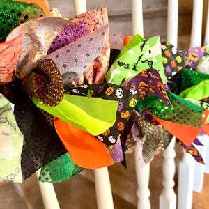 May include: A decorative Halloween garland featuring a collection of fabric strips in various patterns and textures. The strips include prints of ghosts, bats, and pumpkins in orange, green, black, and purple, accented with glittery fabric pieces.
