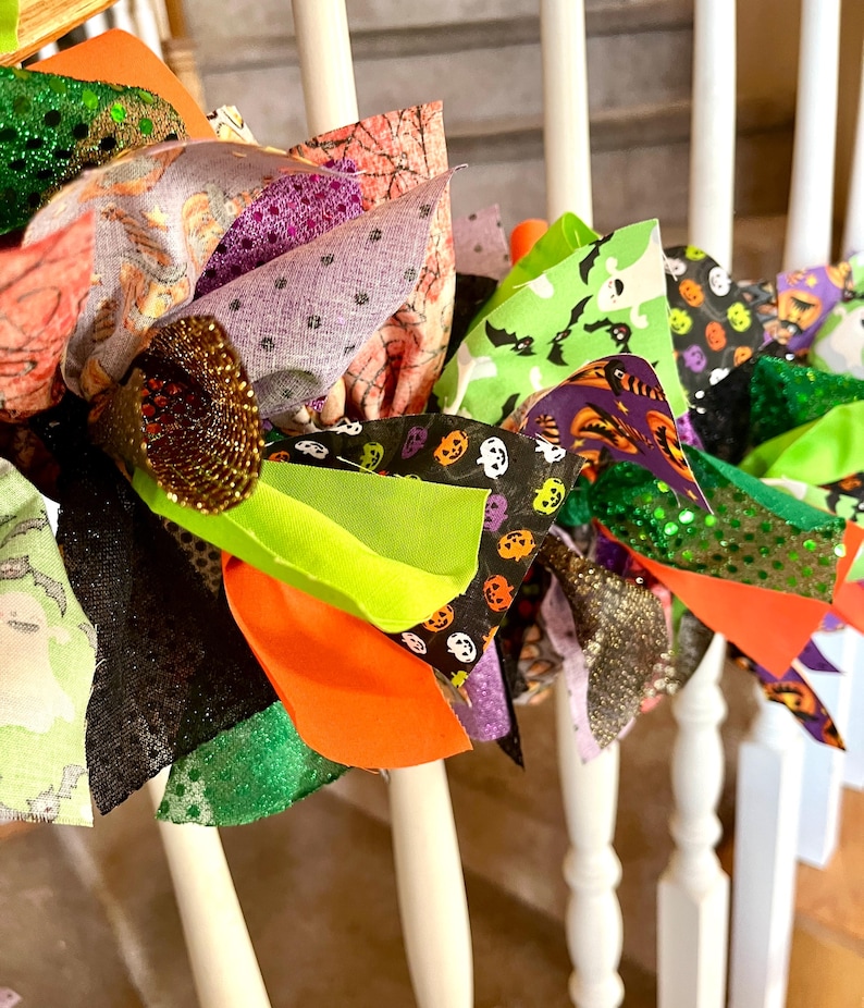 May include: A decorative Halloween garland featuring a collection of fabric strips in various patterns and textures. The strips include prints of ghosts, bats, and pumpkins in orange, green, black, and purple, accented with glittery fabric pieces.