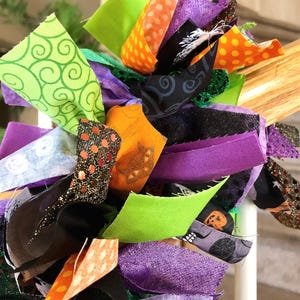 May include: A decorative garland made of numerous fabric strips in shades of purple, orange, lime green, and black. The fabric features various patterns including polka dots, swirls, spider webs, and pumpkin motifs, with some strips having glitter.