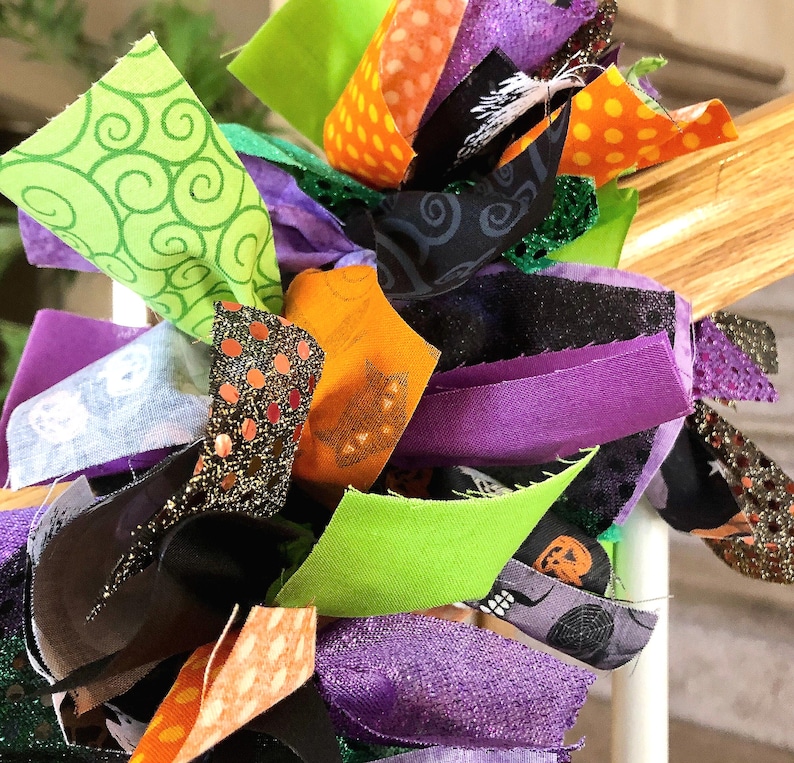 May include: A decorative garland made of numerous fabric strips in shades of purple, orange, lime green, and black. The fabric features various patterns including polka dots, swirls, spider webs, and pumpkin motifs, with some strips having glitter.