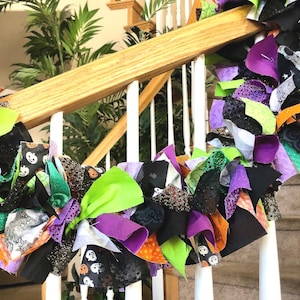 May include: A decorative rag garland featuring a mix of fabric strips in black, bright green, purple, and orange. The fabric includes patterns of pumpkins and abstract designs, with some pieces featuring glittery textures tied along a central cord.