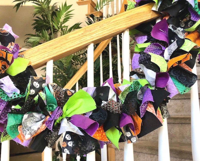 May include: A decorative rag garland featuring a mix of fabric strips in black, bright green, purple, and orange. The fabric includes patterns of pumpkins and abstract designs, with some pieces featuring glittery textures tied along a central cord.