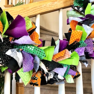 May include: A decorative rag wreath featuring a collection of fabric strips in vibrant shades of orange, lime green, purple, and black. The fabric pieces include various patterns such as polka dots, swirls, and Halloween-themed motifs.
