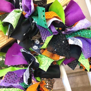 May include: A decorative rag wreath featuring a dense collection of fabric strips in shades of purple, lime green, orange, and black. The fabric includes patterns of jack-o'-lanterns, black cats, and swirling designs, along with glittery textures.