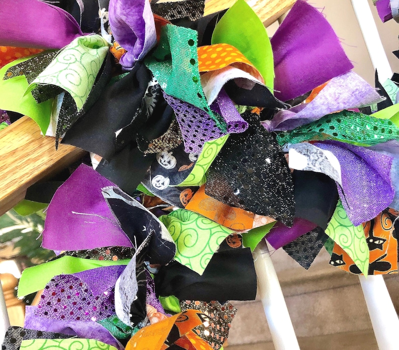 May include: A decorative rag wreath featuring a dense collection of fabric strips in shades of purple, lime green, orange, and black. The fabric includes patterns of jack-o'-lanterns, black cats, and swirling designs, along with glittery textures.