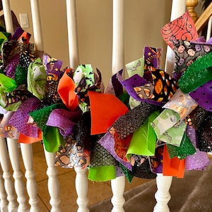 May include: A Halloween-themed rag garland featuring strips of fabric in orange, lime green, purple, and black. The fabric patterns include ghosts, pumpkins, candy corn, and spider webs, tied together to create a festive, textured decorative strand.