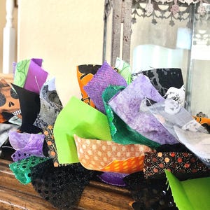May include: A decorative fabric garland featuring a mix of strips in vibrant shades of lime green, purple, orange, and black. The fabric patterns include skulls, polka dots, and solid colors, with some pieces featuring glittery textures and frayed edges.