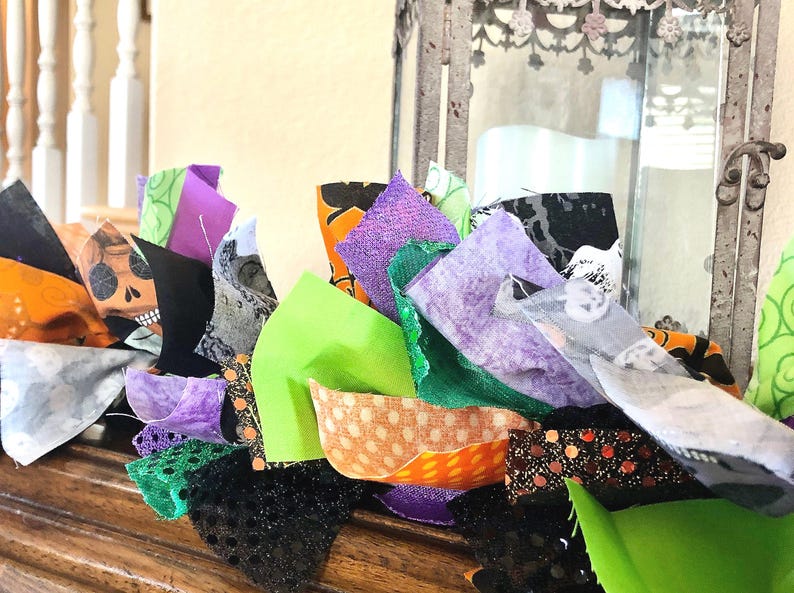 May include: A decorative fabric garland featuring a mix of strips in vibrant shades of lime green, purple, orange, and black. The fabric patterns include skulls, polka dots, and solid colors, with some pieces featuring glittery textures and frayed edges.