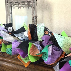 May include: A decorative fabric garland featuring a collection of knotted fabric strips in various patterns and colors including black, purple, bright green, and orange. The fabric pieces display motifs such as pumpkins, swirls, and dots.