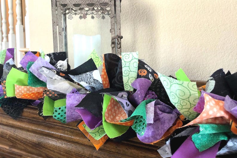 May include: A decorative fabric garland featuring a collection of knotted fabric strips in various patterns and colors including black, purple, bright green, and orange. The fabric pieces display motifs such as pumpkins, swirls, and dots.