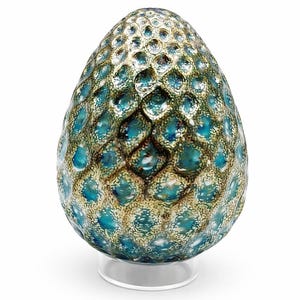 May include: An egg-shaped decorative object with a textured surface. The object is primarily teal and gold, with a pattern of small, raised circles. It sits on a clear, circular stand.