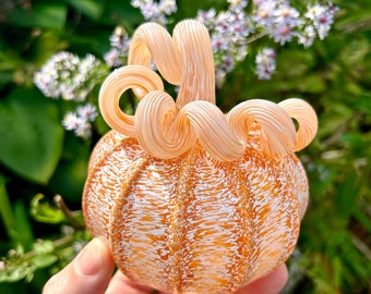 May include: A hand-blown glass pumpkin with a white and orange swirl design. The pumpkin has a textured surface and a curled stem.