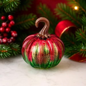 May include: A decorative glass pumpkin with a red, green, and gold striped design. The pumpkin has a brown, curved stem and is set against a backdrop of evergreen branches and red berries, evoking a festive holiday theme.