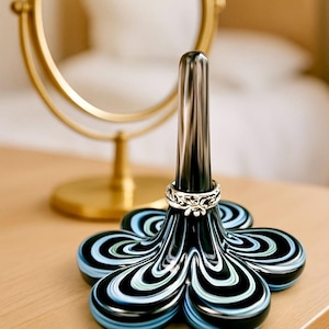 May include: A decorative ring holder with a flower-shaped base. The base features a black, white, and blue swirl pattern. A silver ring sits on the silver post. A gold-colored mirror is in the background.