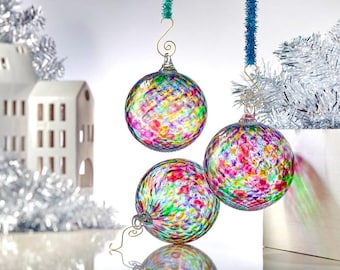 Puede incluir: Tres adornos navideños de cristal de colores con un patrón moteado de rojo, verde, amarillo y azul. Cada adorno está suspendido por un gancho de alambre dorado y una decoración colgante. El fondo incluye una casa de cerámica blanca y oropel plateado.